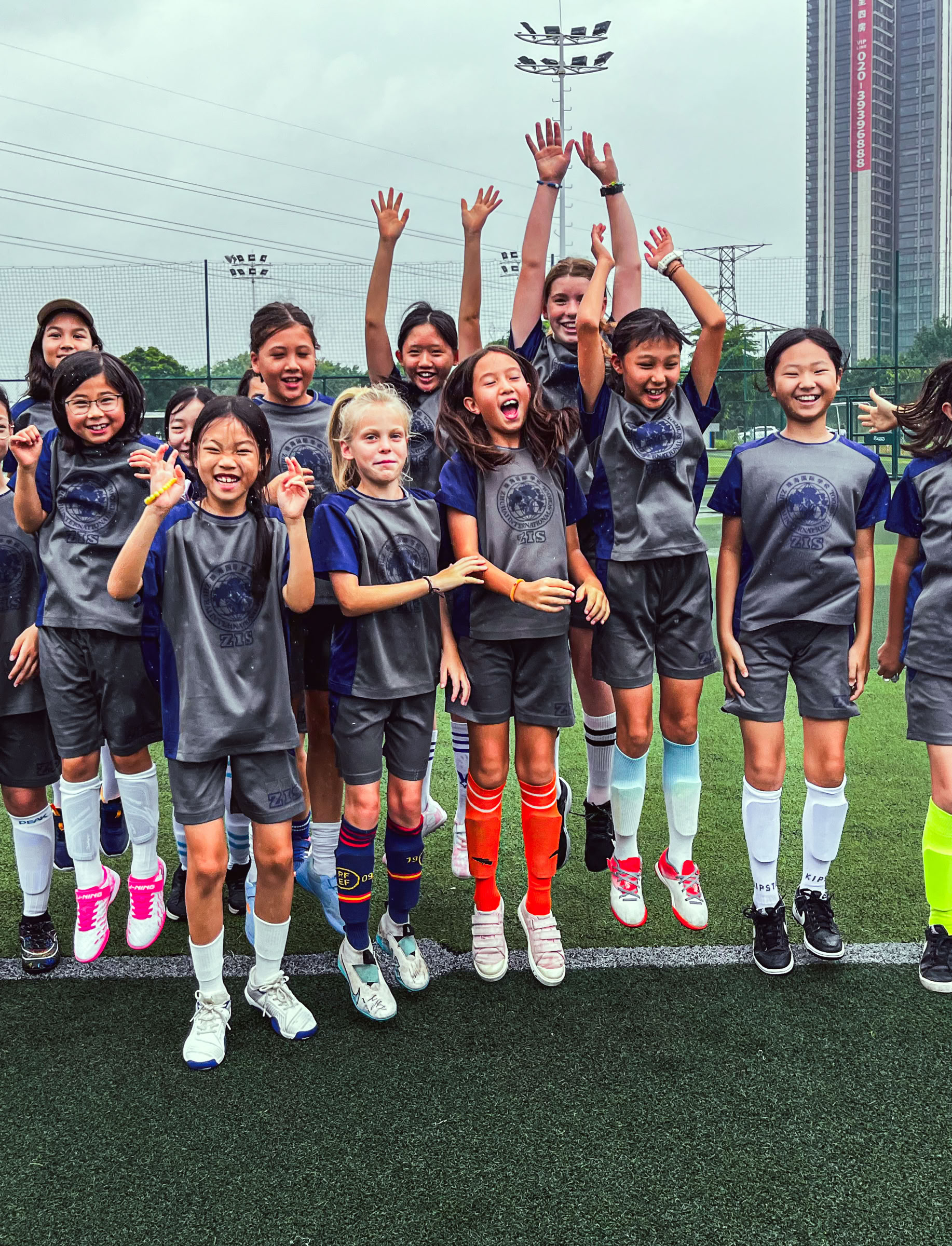 A group of children in soccer uniforms standing on a field, some holding a trophy and others cheering, with buildings and a cloudy sky in the background.