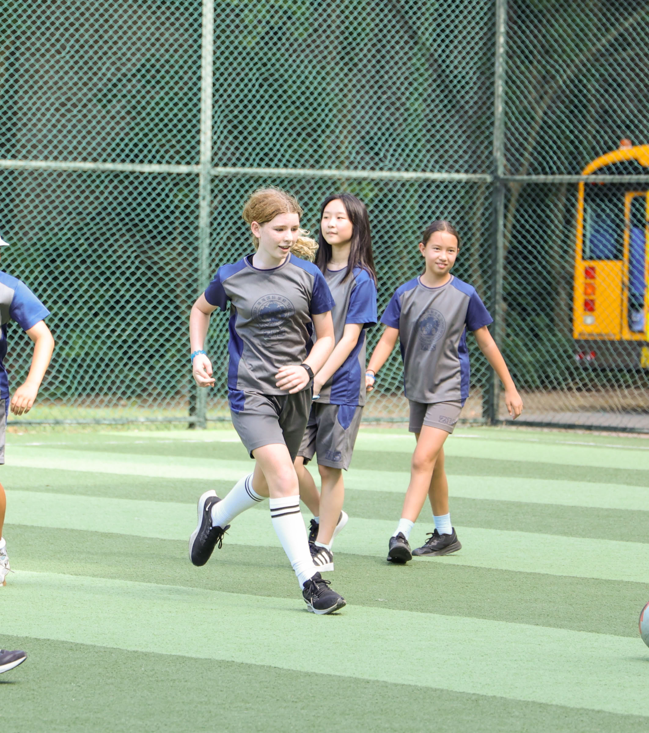 ZIS Students hiking with backpacks walking on a narrow trail through lush green vegetation with mountains in the background under a clear blue sky.